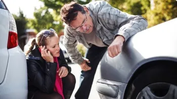 一对夫妇在检查他们银色轿车前部的损坏情况，而女人打电话给她的州立农场代理.